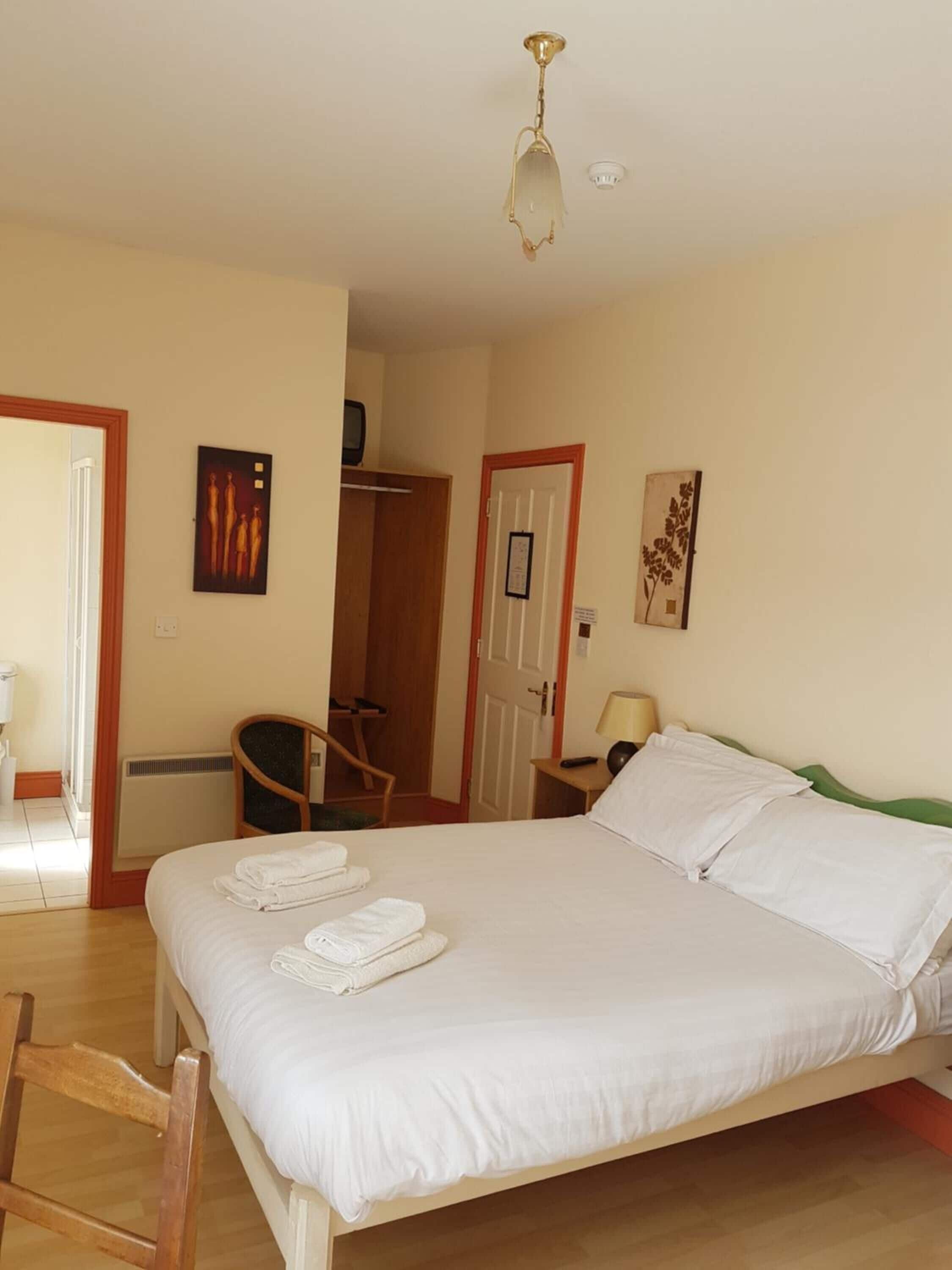 A neatly made bed with white linens and towels, a wooden chair, a small bedside table with a lamp, and a ceiling light fixture.
