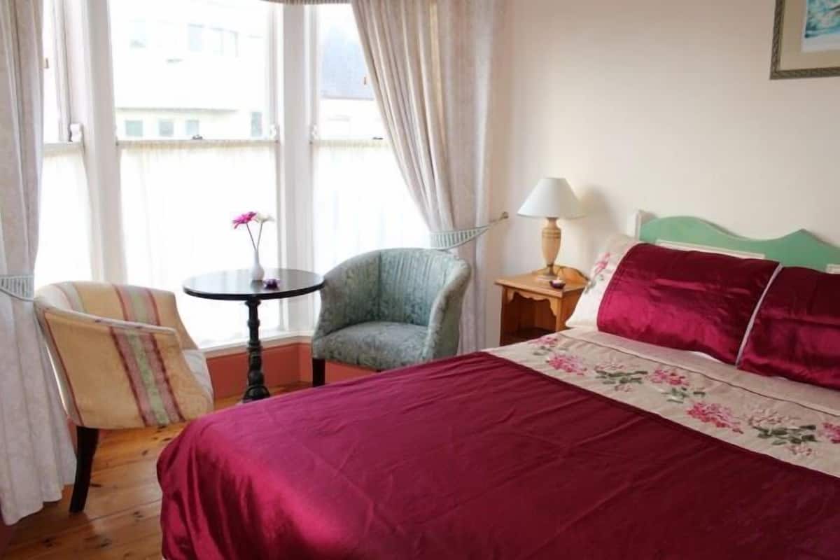 Quarto de hotel com cama, uma cadeira, uma mesinha e uma luminária.