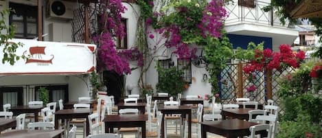Un café al aire libre con mesas y sillas, rodeado de abundante follaje y plantas en flor.