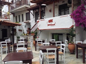 Un café al aire libre con sillas blancas y mesas marrones, rodeado de plantas en macetas y un muro de piedra.