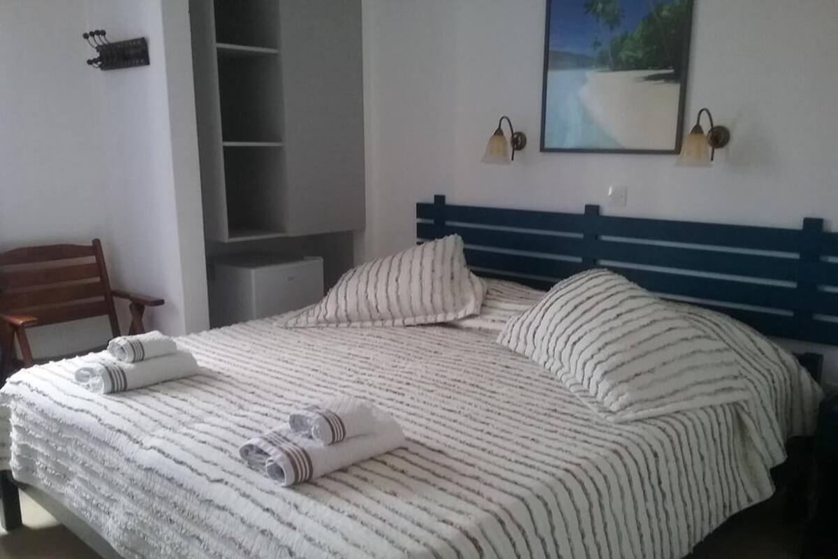 Una cama bien tendida con ropa de cama a rayas y toallas enrolladas, una silla de madera y vista a una playa a través de una ventana.