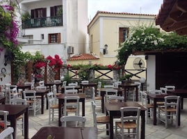 Un café al aire libre con mesas y sillas, rodeado de edificios y plantas florecientes.