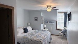 A bedroom with a bed, a ceiling fan, a window with blinds, a chair, and a small table with a lamp. - The Old Lantern Inn and Barn (Charlotte)