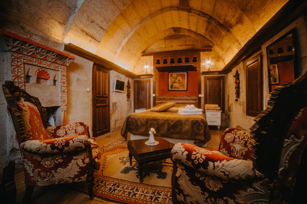 A room with a high arched ceiling, a bed with a red and gold bedspread, two ornate armchairs, a small wooden table, and a patterned rug.
