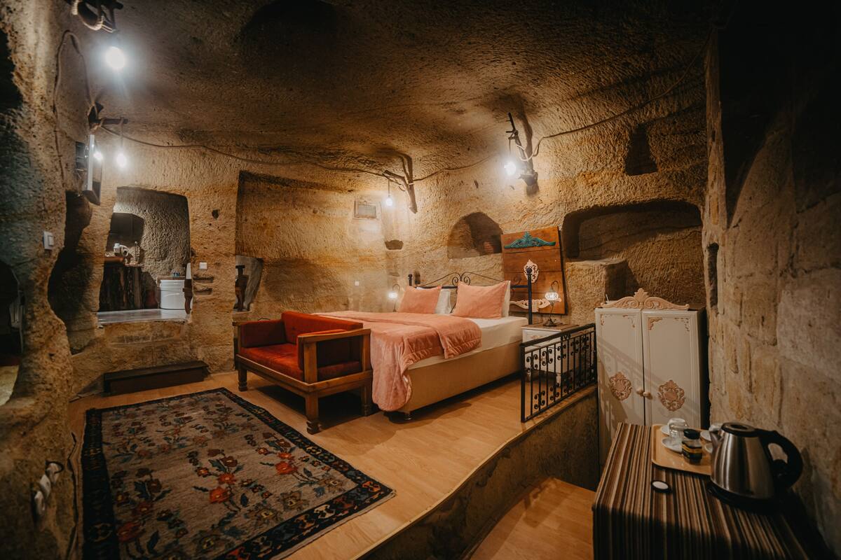A rustic room with a bed, a wooden chair, a small table with a kettle, and a cabinet.
