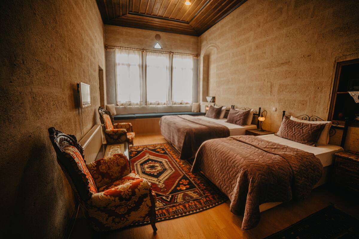 A bedroom with a large bed, a smaller bed, a window with curtains, a patterned rug, and a chair.