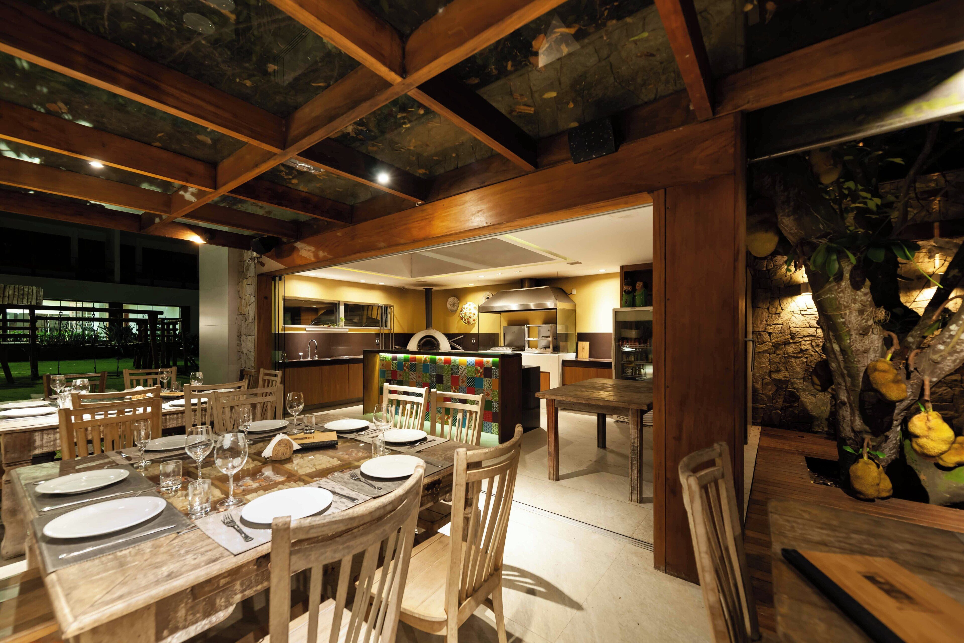 A dining area with wooden tables and chairs, a kitchen in the background, and a stone wall with hanging fruits.