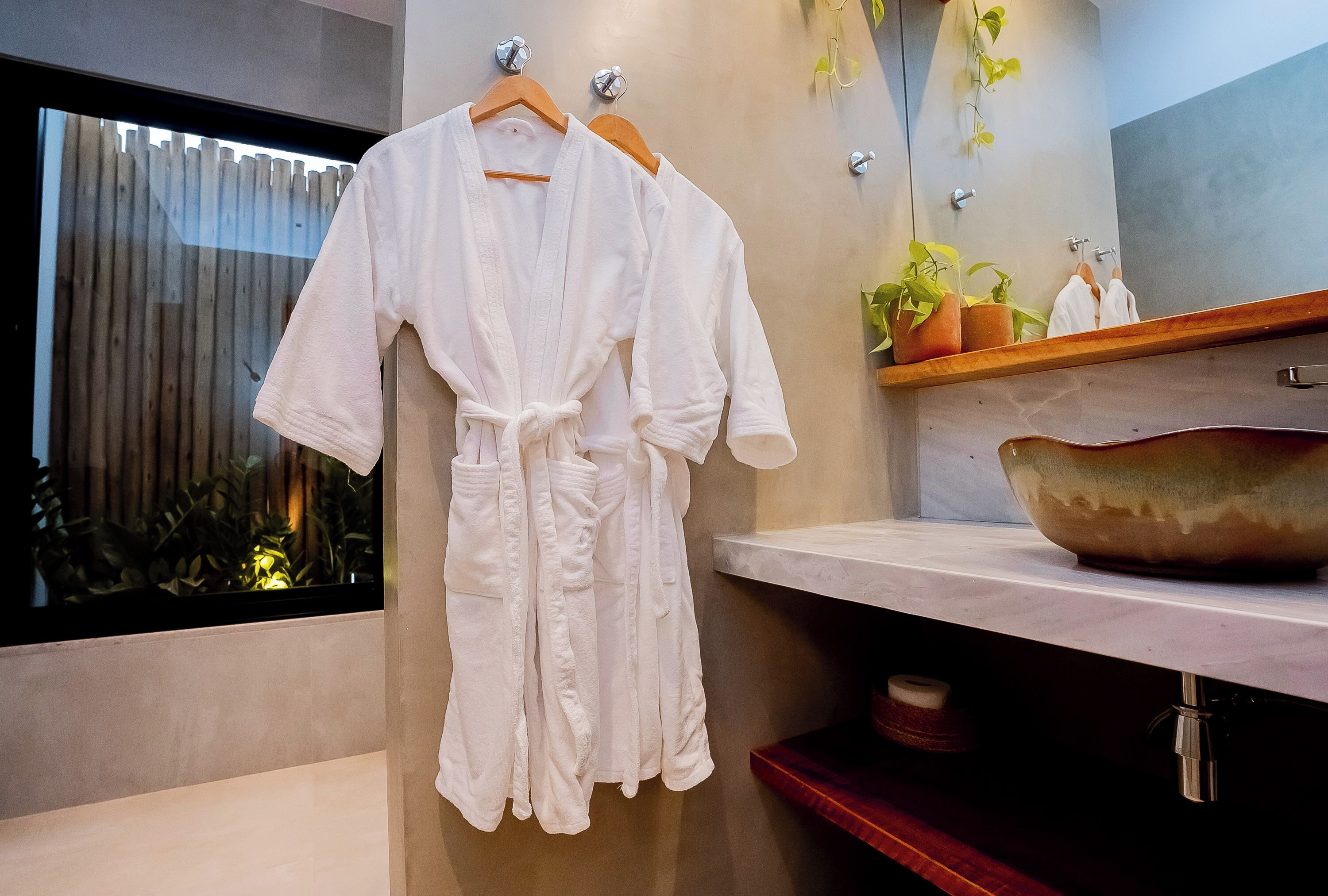 A white robe hanging on a wooden hanger in a modern bathroom with a large mirror, a wooden shelf with plants, and a large ceramic bowl.