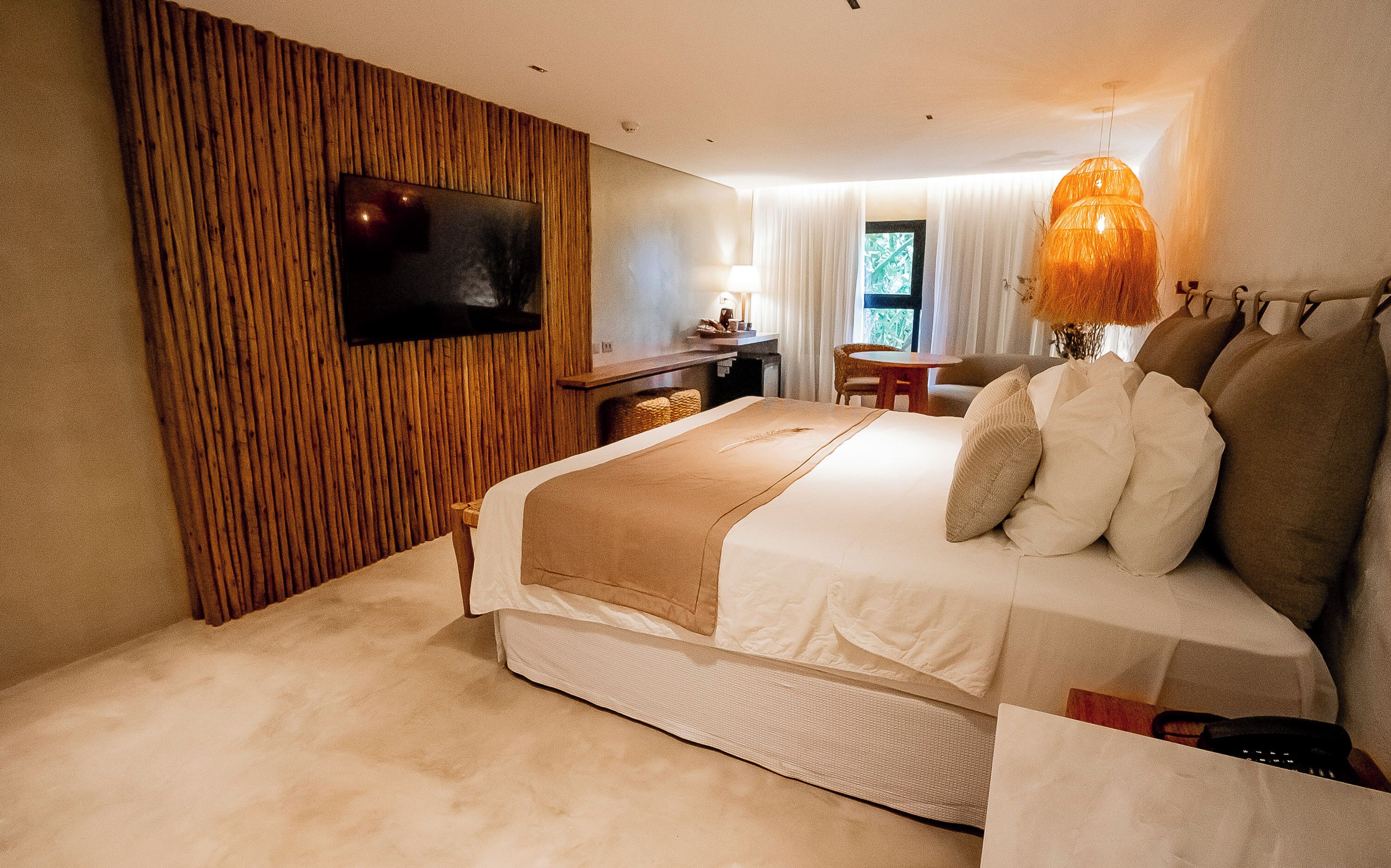 A hotel room with a large bed, a flat-screen TV mounted on the wall, a desk with a lamp, and a view of the outdoors through the window.