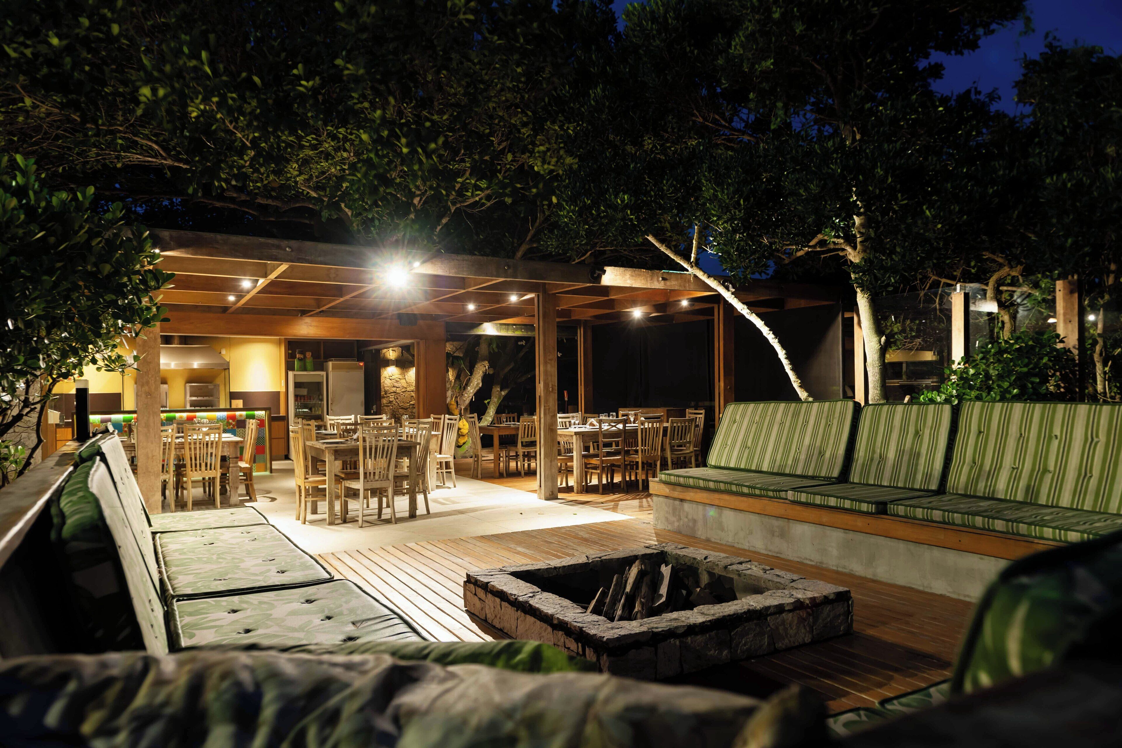 An outdoor dining area with wooden tables and chairs, a fire pit, and a covered seating area with cushions.