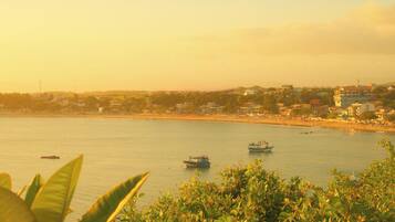 Vista costera con barcos en el agua, una playa y un paisaje urbano al fondo.