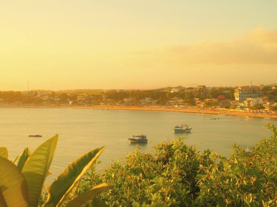 Vista costera con barcos en el agua, una playa y un paisaje urbano al fondo.