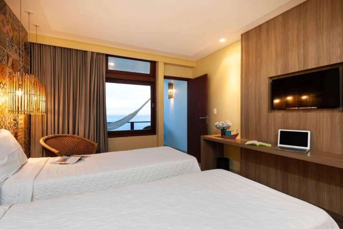 A hotel room with two beds, a TV, a laptop, and a view of the sea through a large window.