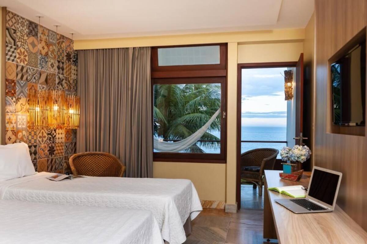 A hotel room with a bed, a desk with a laptop, a chair, a large window with a view of palm trees and the sea, and a decorative wall with patterned tiles.