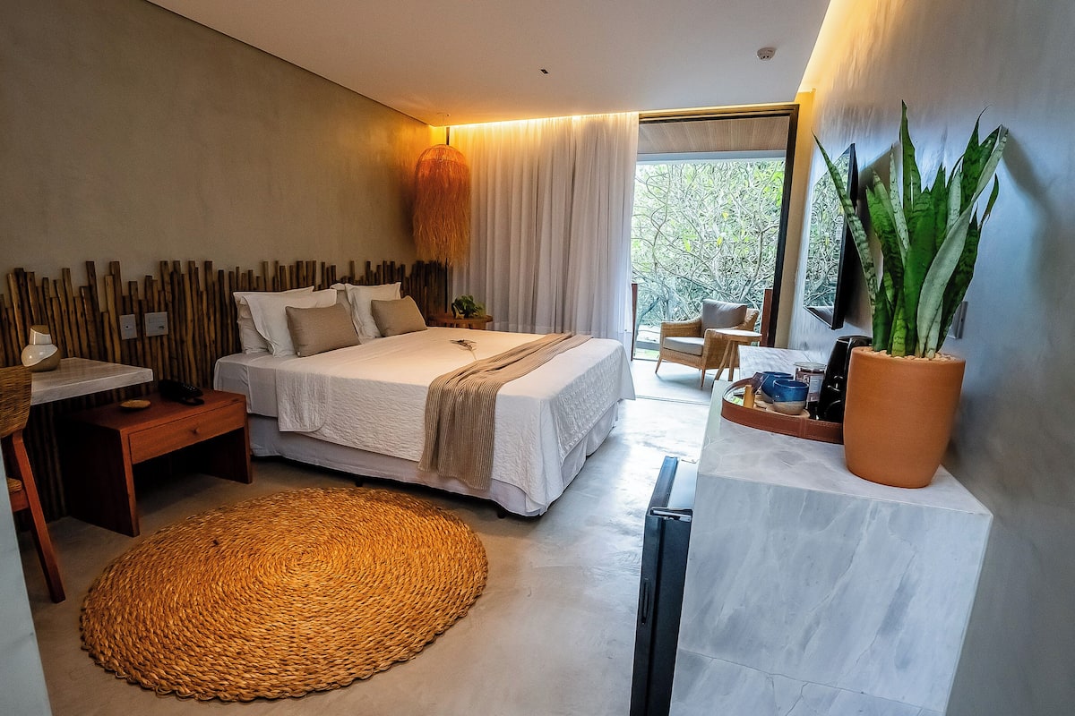 A modern bedroom with a large bed, bedside tables, a woven rug, a potted plant, and a view of greenery outside.
