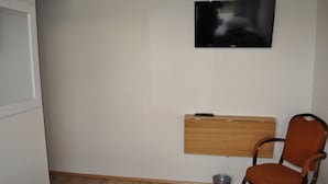 Una habitación con una televisión montada en la pared, un estante de madera, una silla y un cubo de basura.