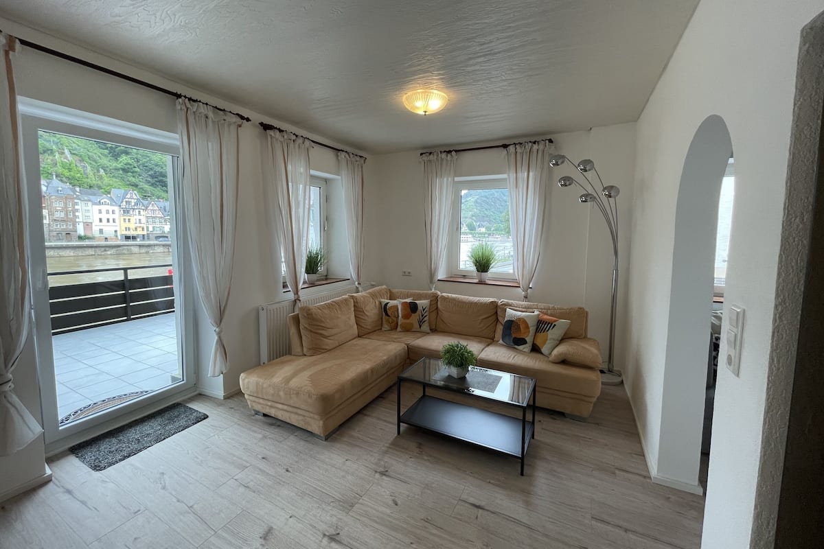 Una sala de estar moderna con un sofá seccional color beige, una mesa de centro de vidrio y vistas a un río y edificios a través de amplios ventanales.