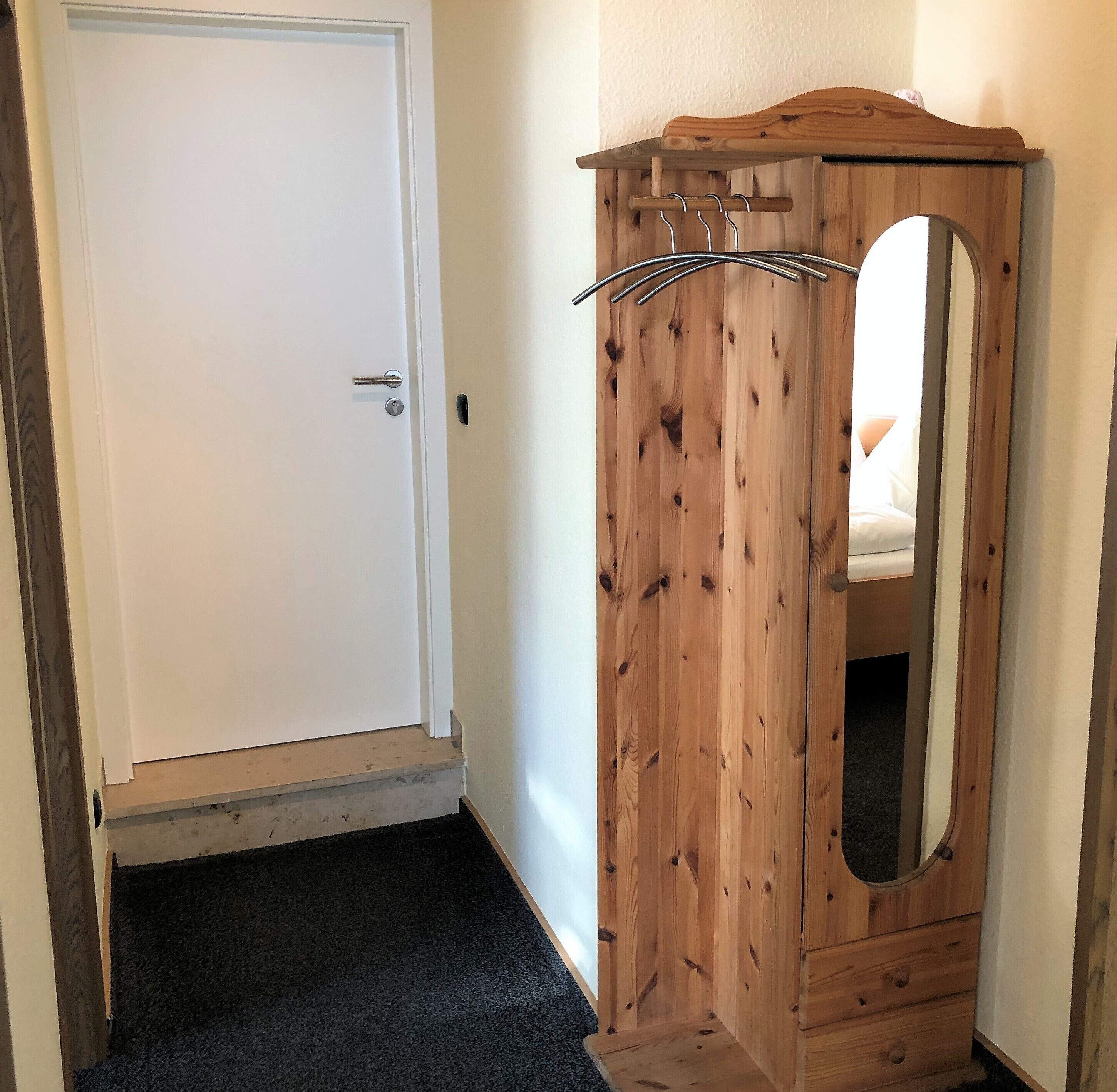 A wooden wardrobe with a mirror and a coat rack, a white door, and a dark carpeted floor.