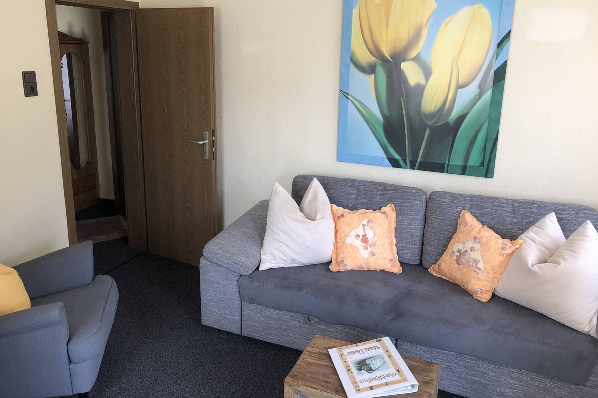 Un salón con un sofá gris, una mesita de madera y un cuadro de tulipanes amarillos en la pared.