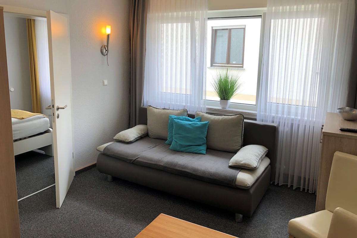 Habitación de hotel con sofá, una mesita, planta en maceta y ventana con cortinas translúcidas.