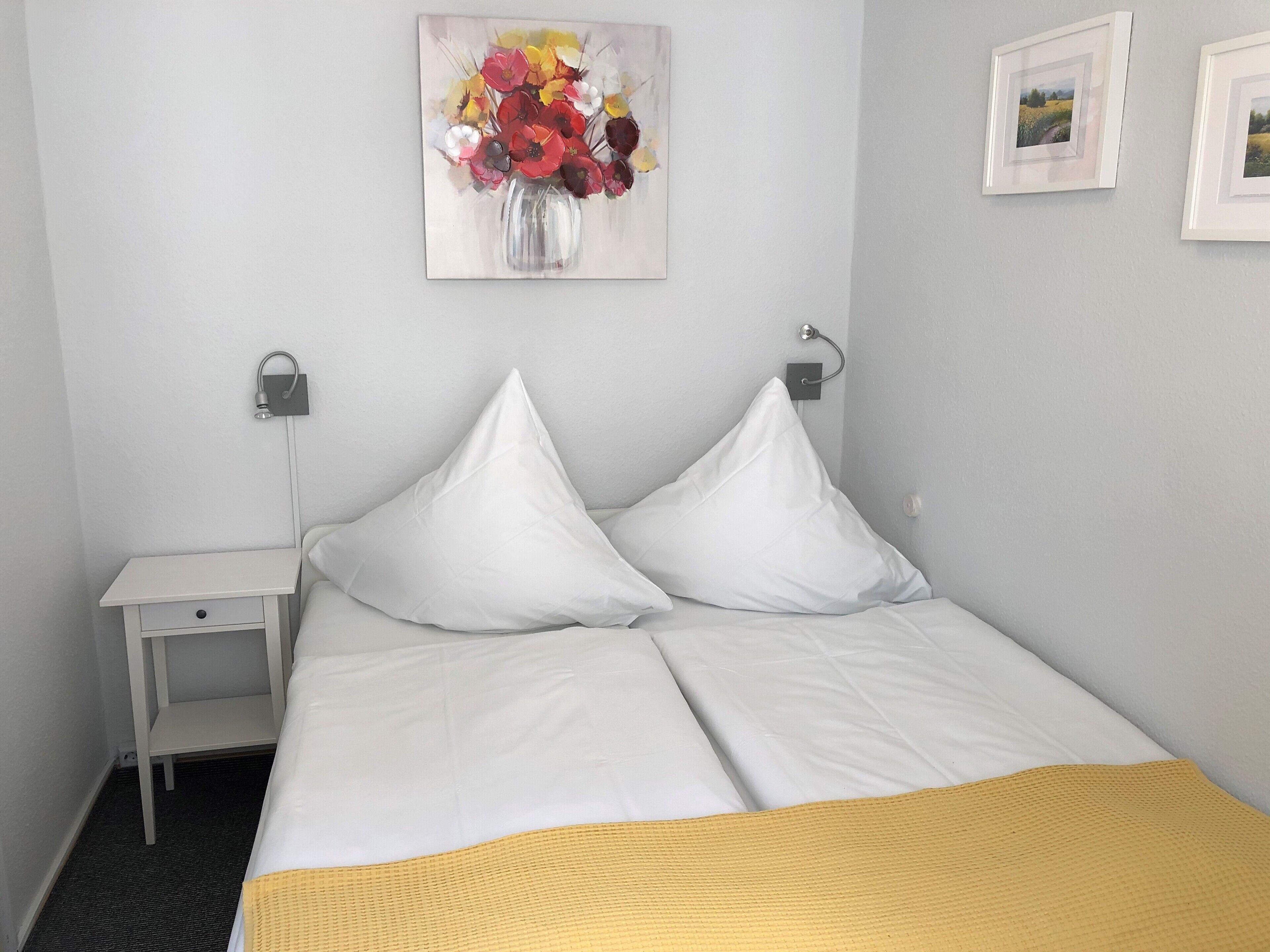 A hotel room with a bed, a nightstand, a lamp, a painting, and a small shelf.