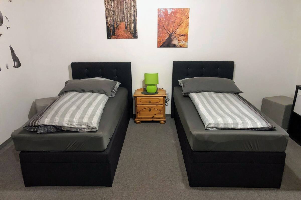 A room with two single beds, a small wooden nightstand with a green lamp, and two framed pictures on the wall.