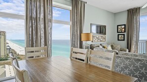 A room with a wooden dining table, chairs, a sofa with patterned cushions, a lamp, and a view of the beach and ocean.