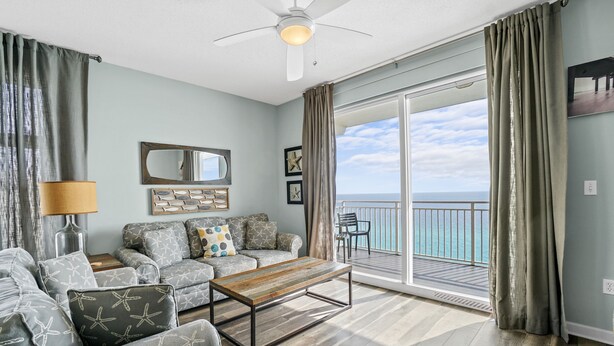A living room with a sofa set, a coffee table, a ceiling fan, and a view of the ocean through a sliding glass door.