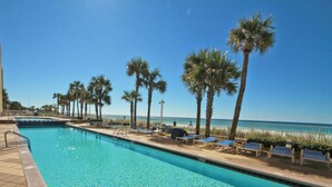 A swimming pool with clear blue water, surrounded by palm trees and lounge chairs, with a beach and ocean in the background.