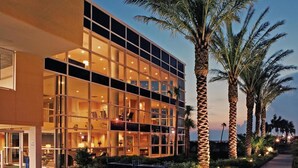 A modern hotel with a glass facade, palm trees, and a well-lit entrance.