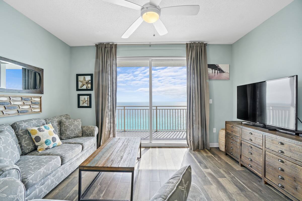 A living room with a sofa, coffee table, TV, and a balcony with a view of the ocean.