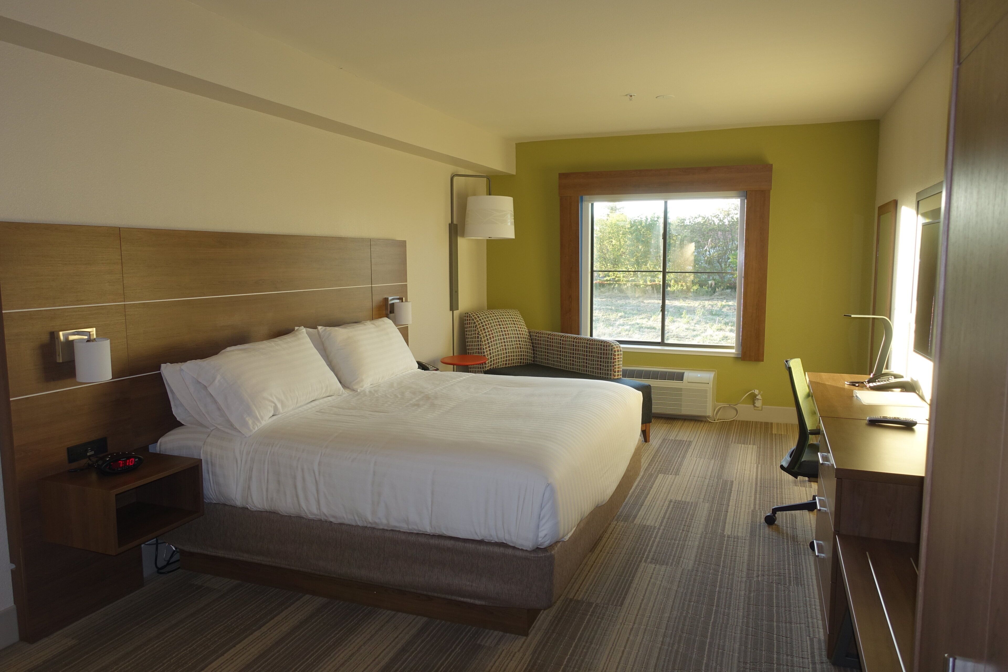 Una habitación de hotel con una cama grande, mesitas de noche, un escritorio con silla, una ventana y una lámpara de pared.