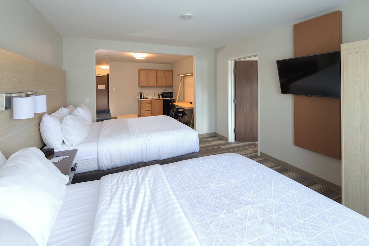A hotel room with two beds, a TV, a desk, and a kitchenette.
