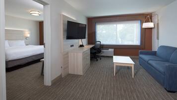 Habitación de hotel con cama, escritorio con computadora, televisor, ventana con vista a árboles y sofá azul.