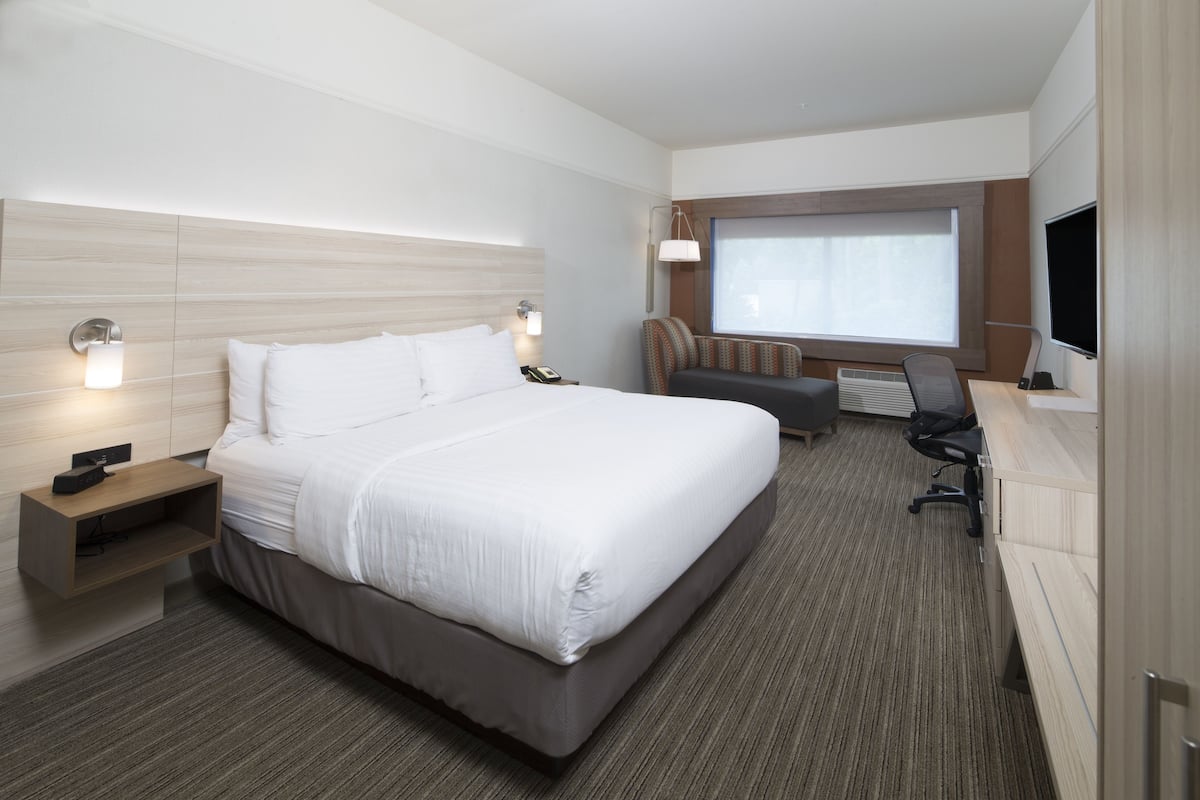 Ein modernes Hotelzimmer mit einem großen Bett, einem Schreibtisch mit Stuhl, einem Fernseher und einem Fenster mit Jalousien.