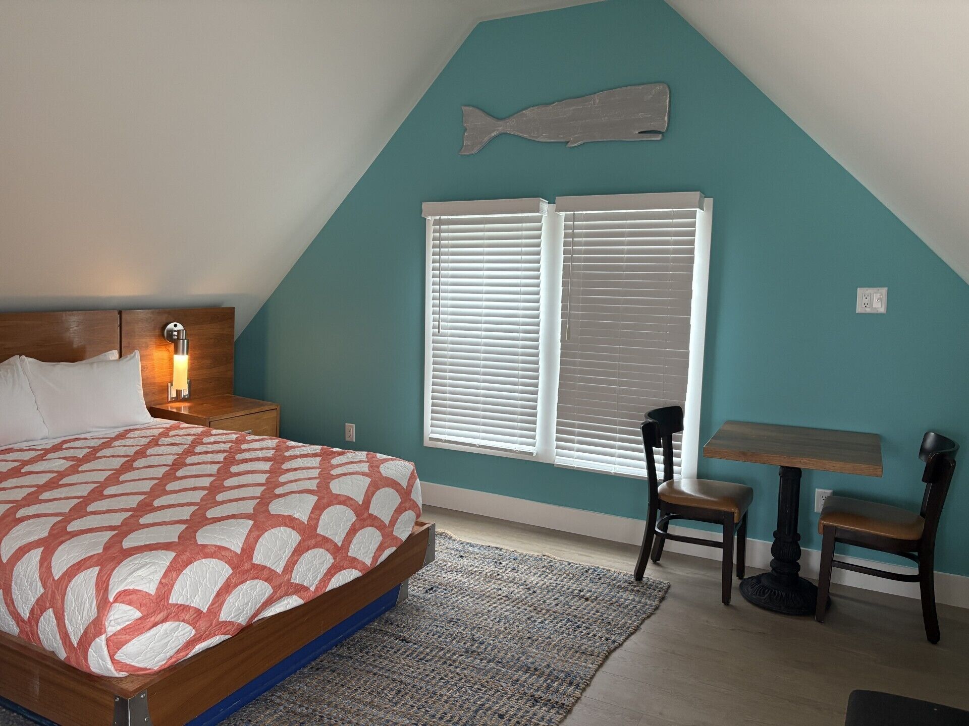 A bedroom with a bed, bedside table, lamp, window with blinds, and a wall-mounted fish decoration.