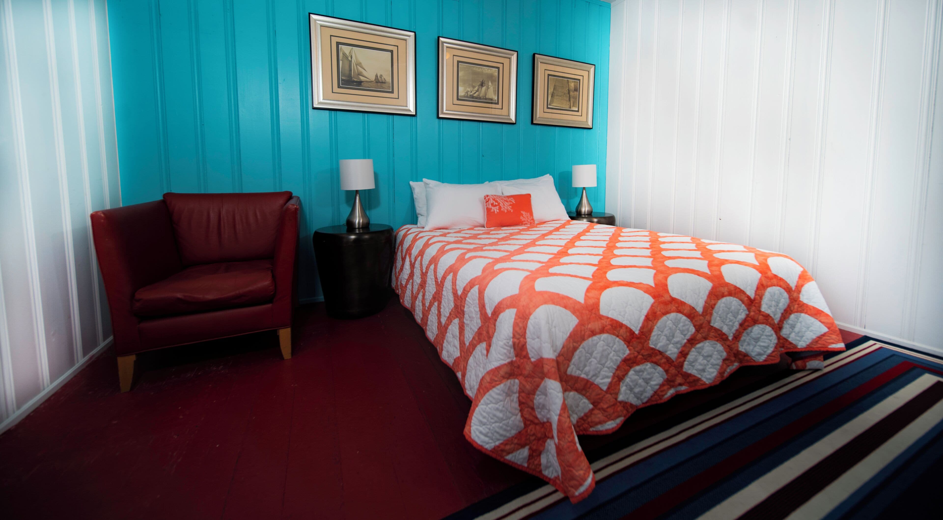 A bedroom with a red leather chair, a bed with an orange and white patterned bedspread, a nightstand with a lamp, and three framed pictures on the wall.