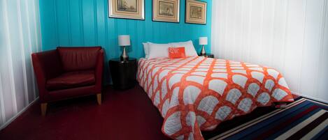 A bedroom with a red leather chair, a bed with an orange and white patterned bedspread, a nightstand with a lamp, and three framed pictures on the wall.