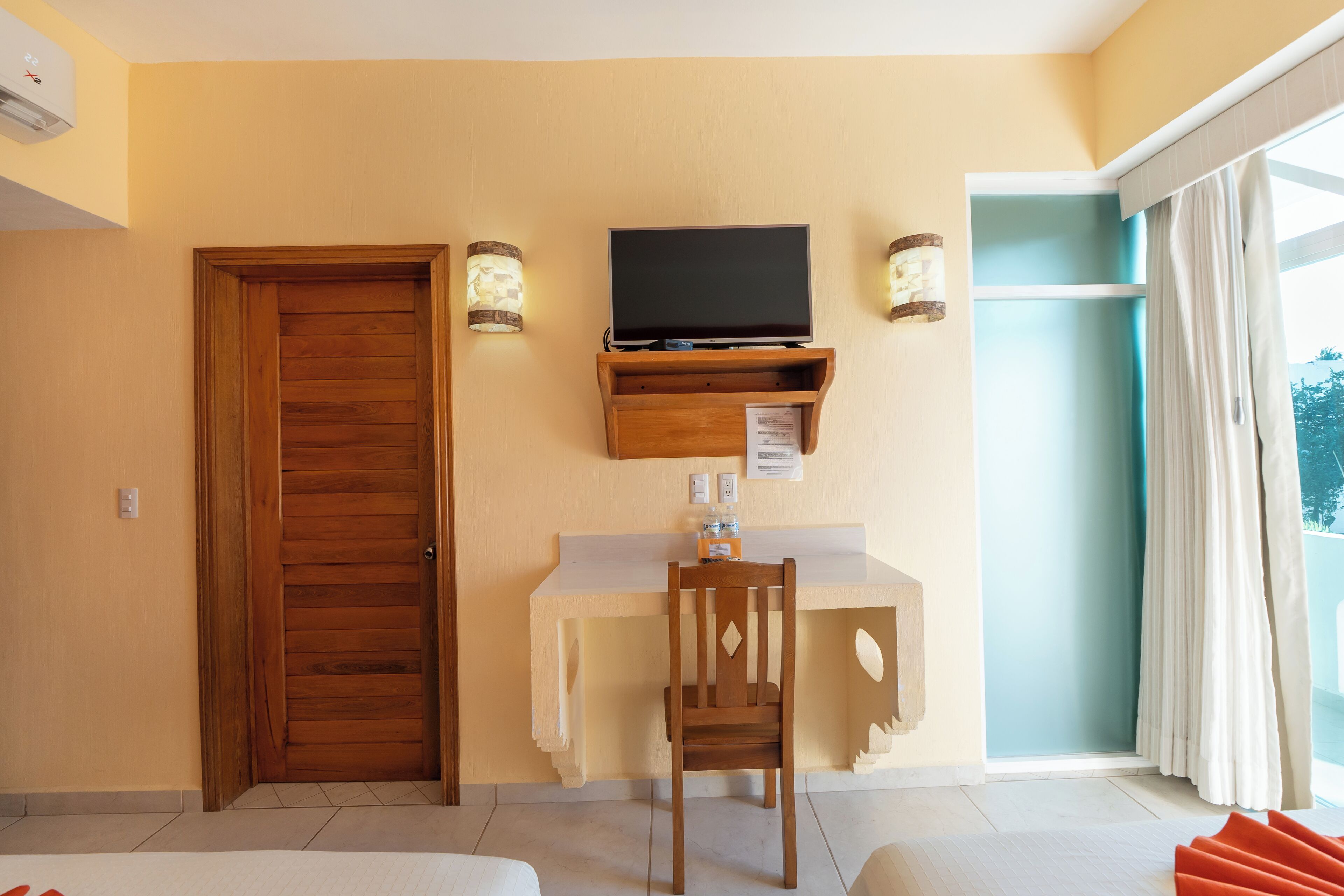 Une chambre d’hôtel avec une porte en bois, un bureau avec une télévision, deux appliques murales et une grande fenêtre avec des rideaux.