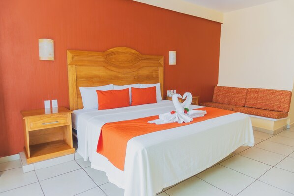 A hotel room with a wooden headboard, a bed with white and orange bedding, a nightstand, and a sofa with orange cushions.