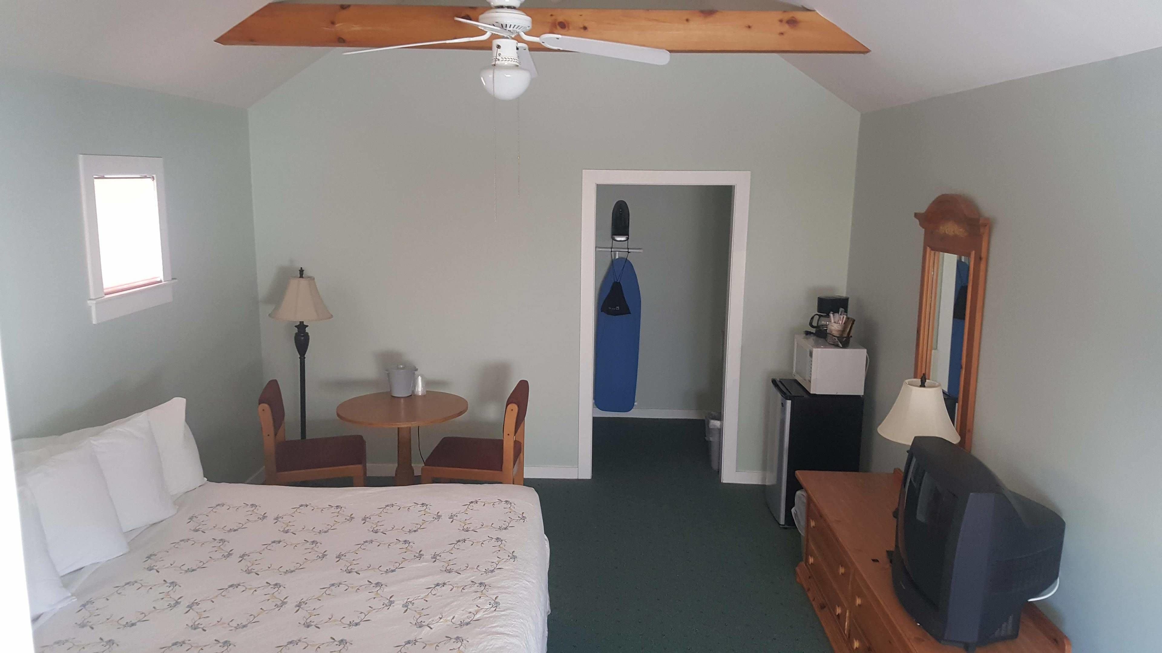 A bedroom with a bed, a desk, a chair, a ceiling fan, a mirror, and a door leading to a bathroom.