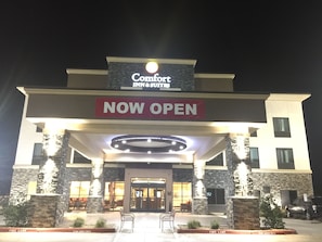 Entrada d'un hotel Comfort Inn & Suites amb un cartell que diu 'NOW OPEN', pilars de pedra i seients a l'exterior.
