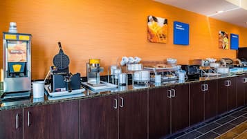 Una barra de desayuno con diversas opciones de comida y bebida, incluyendo una máquina para gofres, cafeteras y un dispensador de bebidas autoservicio.