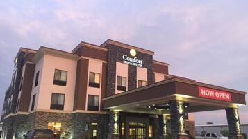 A Comfort Inn & Suites hotel with a 'NOW OPEN' sign, a covered entrance, and a parking area with vehicles.