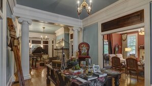 A spacious living room with a grand chandelier, a piano, and a large table with books and decorative items. - The English Merchant's Inn (Waxahachie)