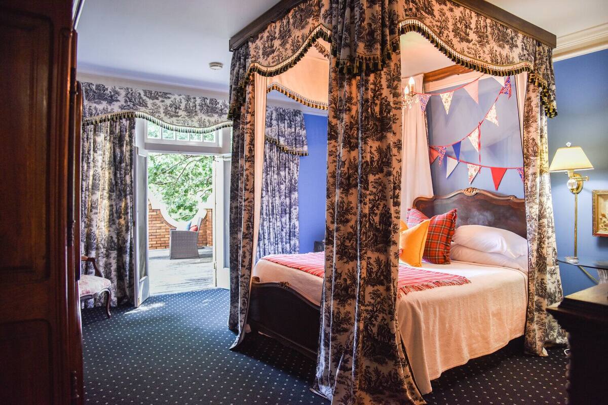 A four-poster canopy bed with a view to a garden, a wooden chair, and a lamp on a bedside table.