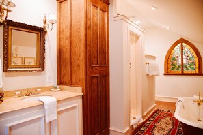 A bathroom with a wooden vanity, a large mirror, a bathtub, and a stained glass window. - The English Merchant's Inn (Waxahachie)