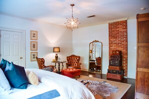 A bedroom with a bed, two chairs, a table with a lamp, a mirror, and a brick wall. - The English Merchant's Inn (Waxahachie)