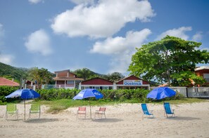 A beach with four beach chairs and two blue umbrellas, a building with a sign reading "Pousada Oceánica", and a white fence in the background. - Pousada Oceânica (Bombinhas)