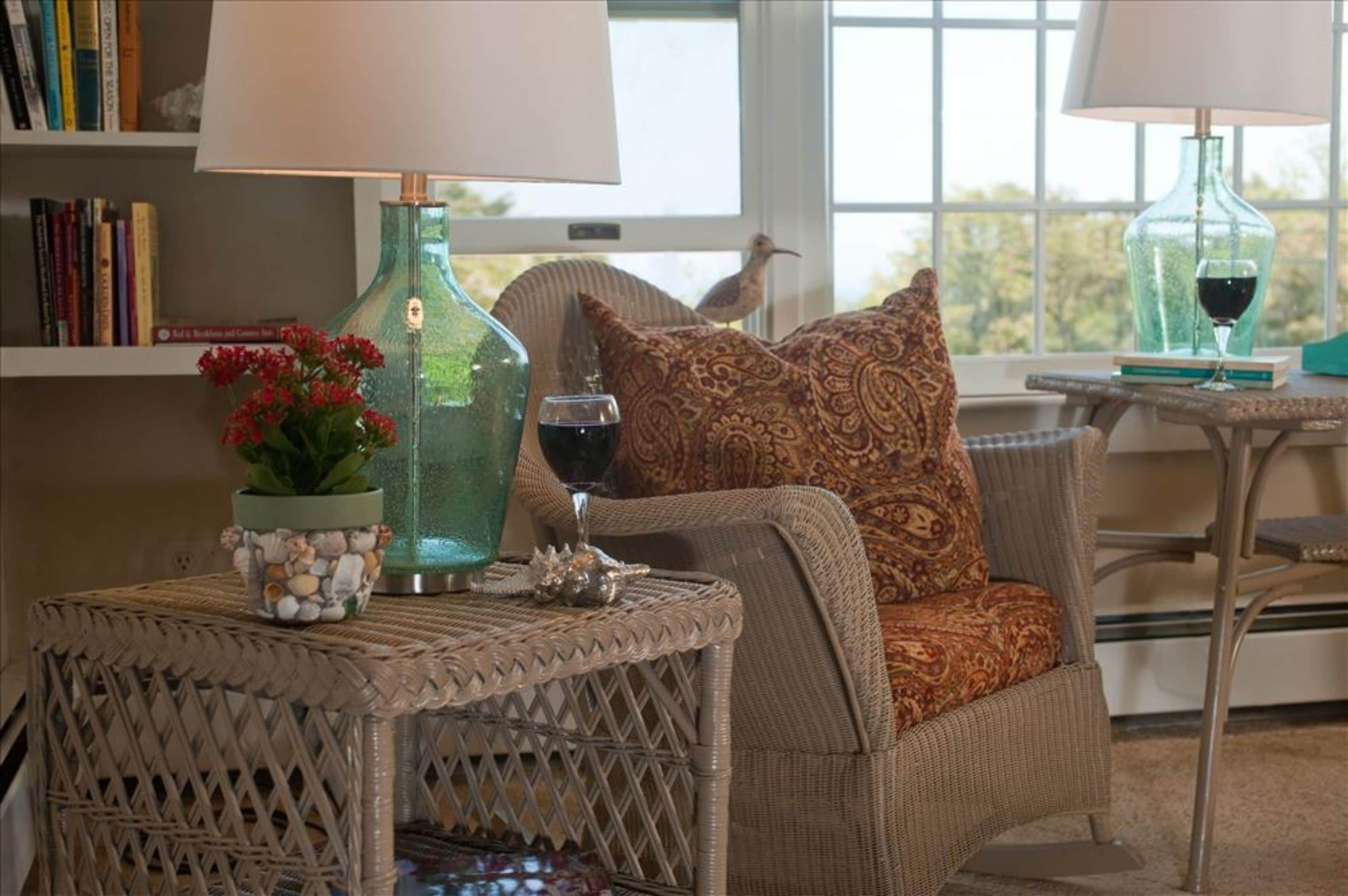 A cozy living room with wicker furniture, a patterned cushion, a vase with red flowers, and a bookshelf with books.